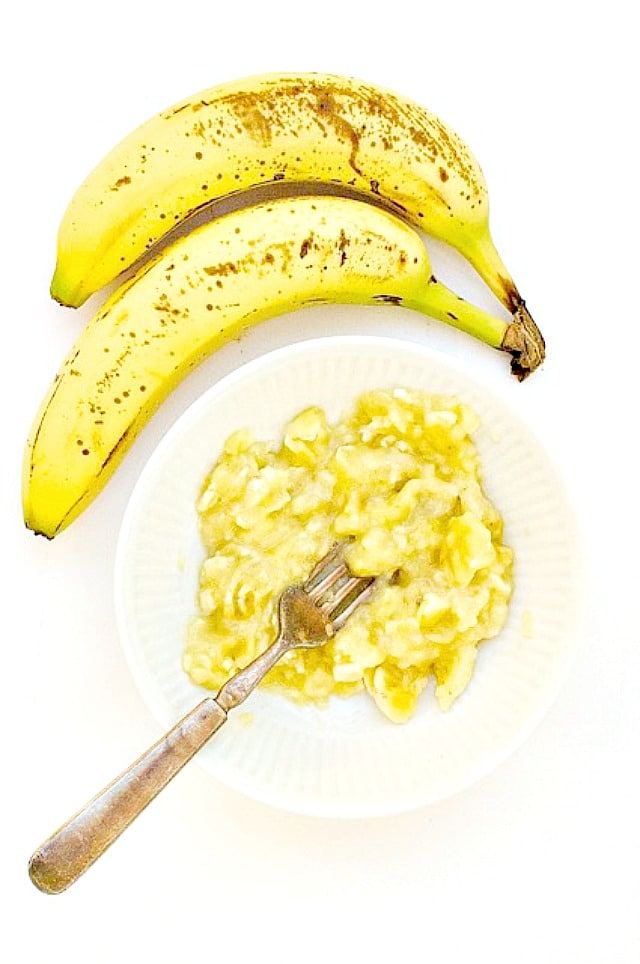A bowl of mashed bananas sits below two very ripe bananas.