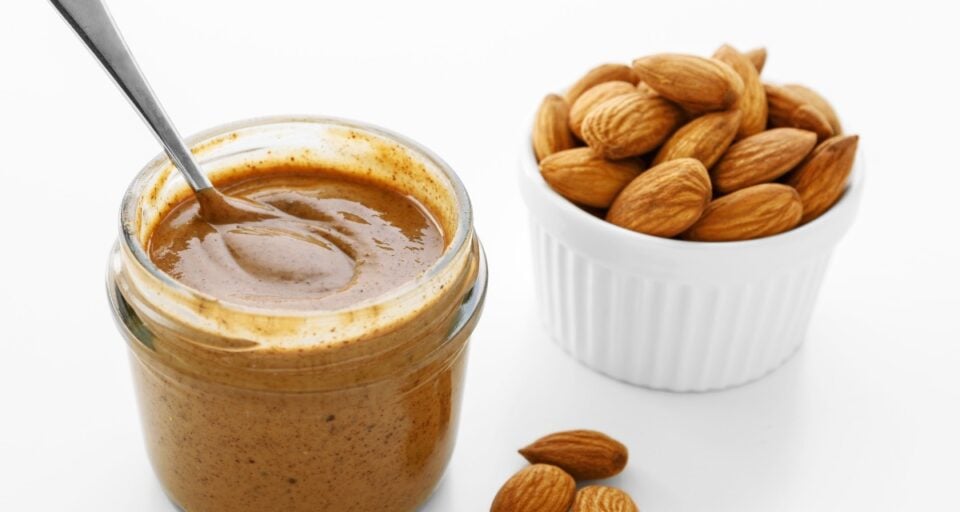 A jar of almond butter has a spoon it. There is a bowl of almonds beside it along with a few almonds on the table.