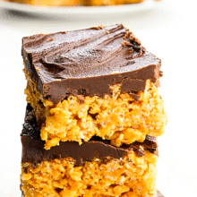 There's a stack of three Rice Krispies treats with chocolate fudge on top on a white counter. In the background is a plate with more of the treats. The text at the top of the image reads, Chocolate Peanut Butter Rice Krispie Treats.