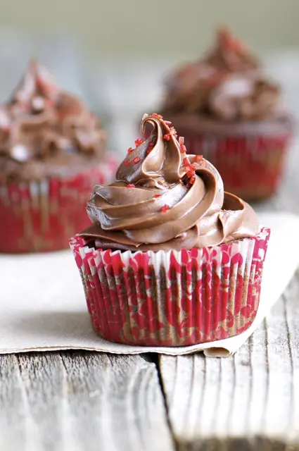 A chocolate cherry cupcake with a large swirl of chocolate frosting on top sits on a wooden table. There are two more cupcakes behind it.