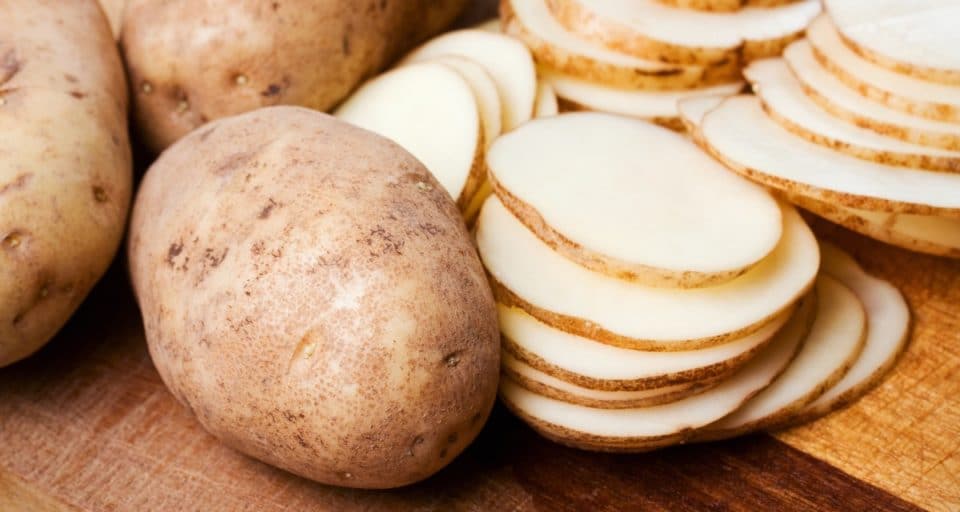 Russet potatoes sit on a cutting board, with several sliced potatoes sitting beside them.
