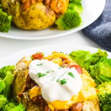 A baked potato sits on a plate with broccoli around it. The potato has lots of toppings like chili and sour cream. There's another potato just like it in the background sitting by a blue kitchen towel. The text at the top of the page reads, Loaded Vegan Baked Potatoes.