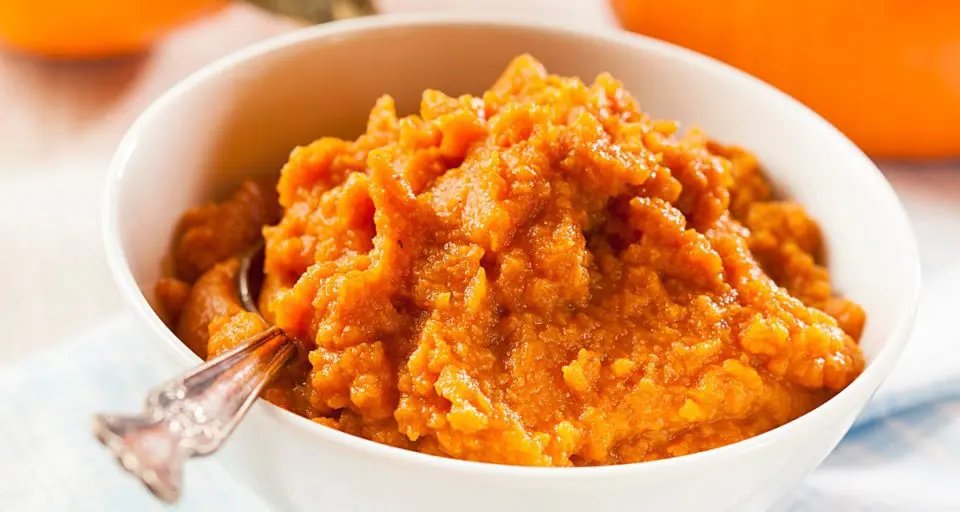 A bowl of pumpkin puree has a spoon it. There is a pumpkin barely visible in the background.