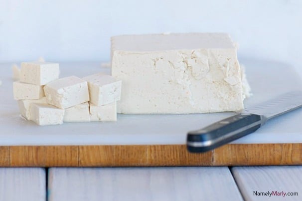 A block of tofu sits on a cutting board next to a knife and several smaller cubes of tofu.