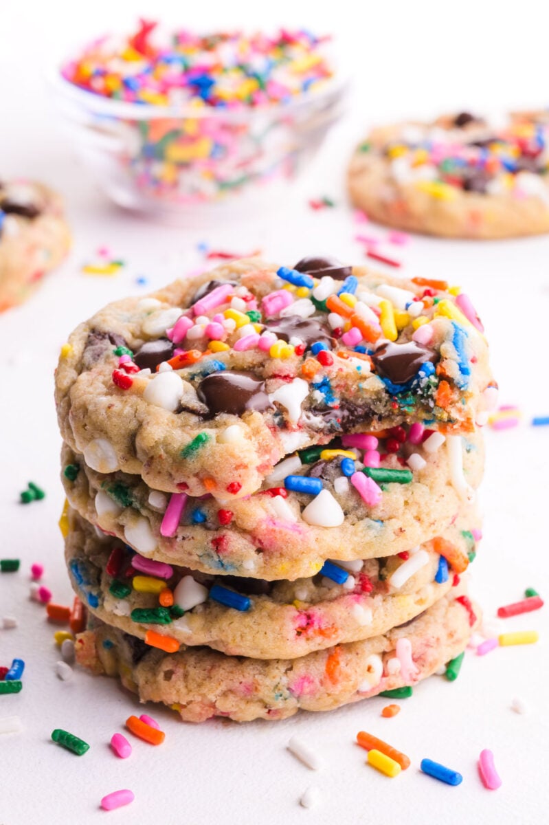 A stack of Vegan Chocolate Chip Sprinkle Cookies. The top cookie has a bite taken out and colorful sprinkles are scattered around.