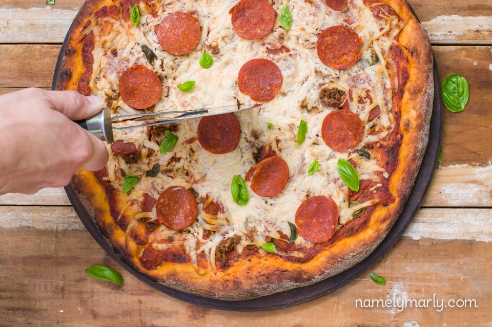 A hand reaches in with the pizza cutter to cut the pepperoni pizza.