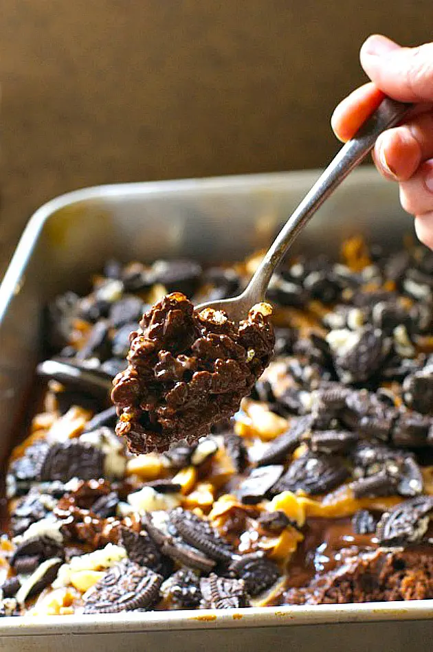 A hand holds a spoon full of chocolate rice crispy mixture hovering over a pan of layered brownies.