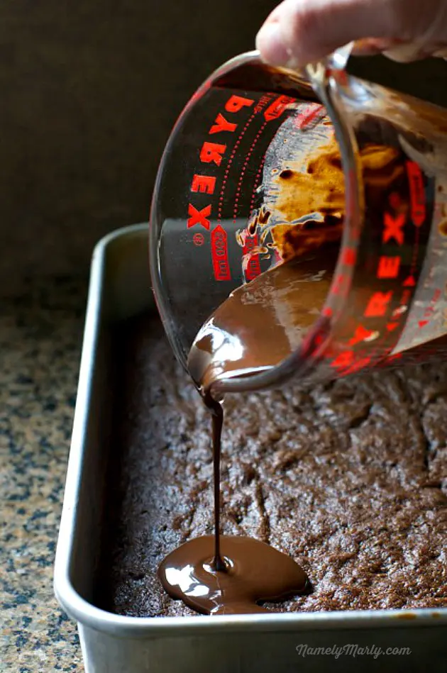 A pyrex measuring cup is full of chocolate sauce that is being poured over a pan of brownies.