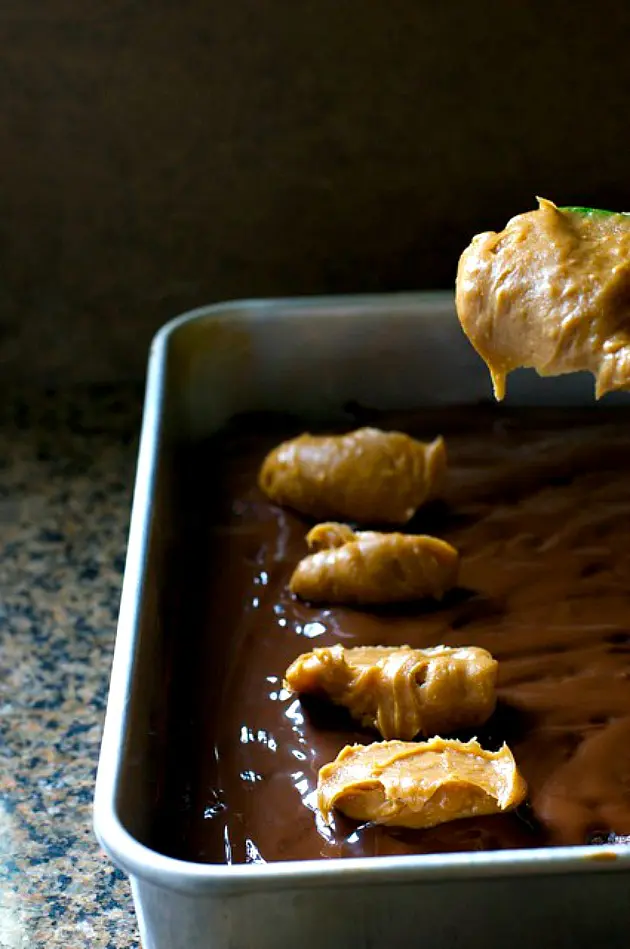A spoon is being used to drop dollops of peanut butter sauce over brownies in a pan.