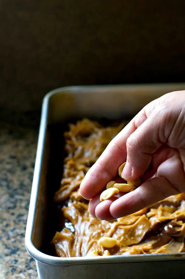 A hand holds peanuts, distributing them over a pan full of layered brownies.