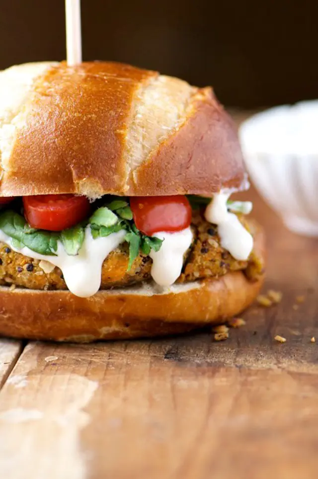 A closeup of a Sweet Potato Veggie Burger with vegan mayo drizzled over the edges, greens and chopped tomatoes.
