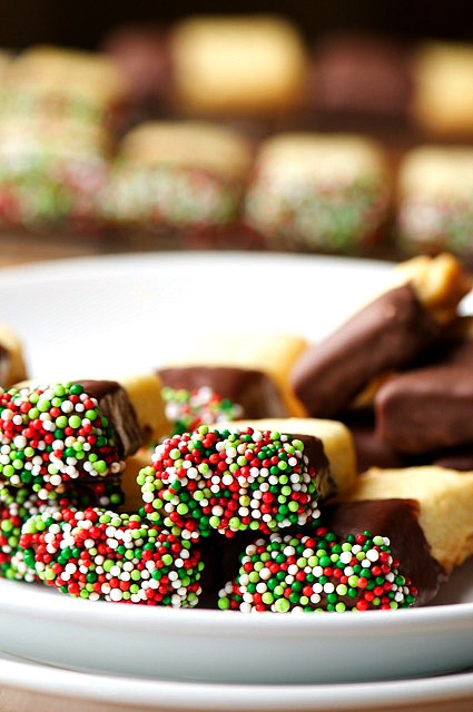 Several shortbread cookies dipped in chocolate and sprinkles are sitting on a plate with more cookies behind it. 