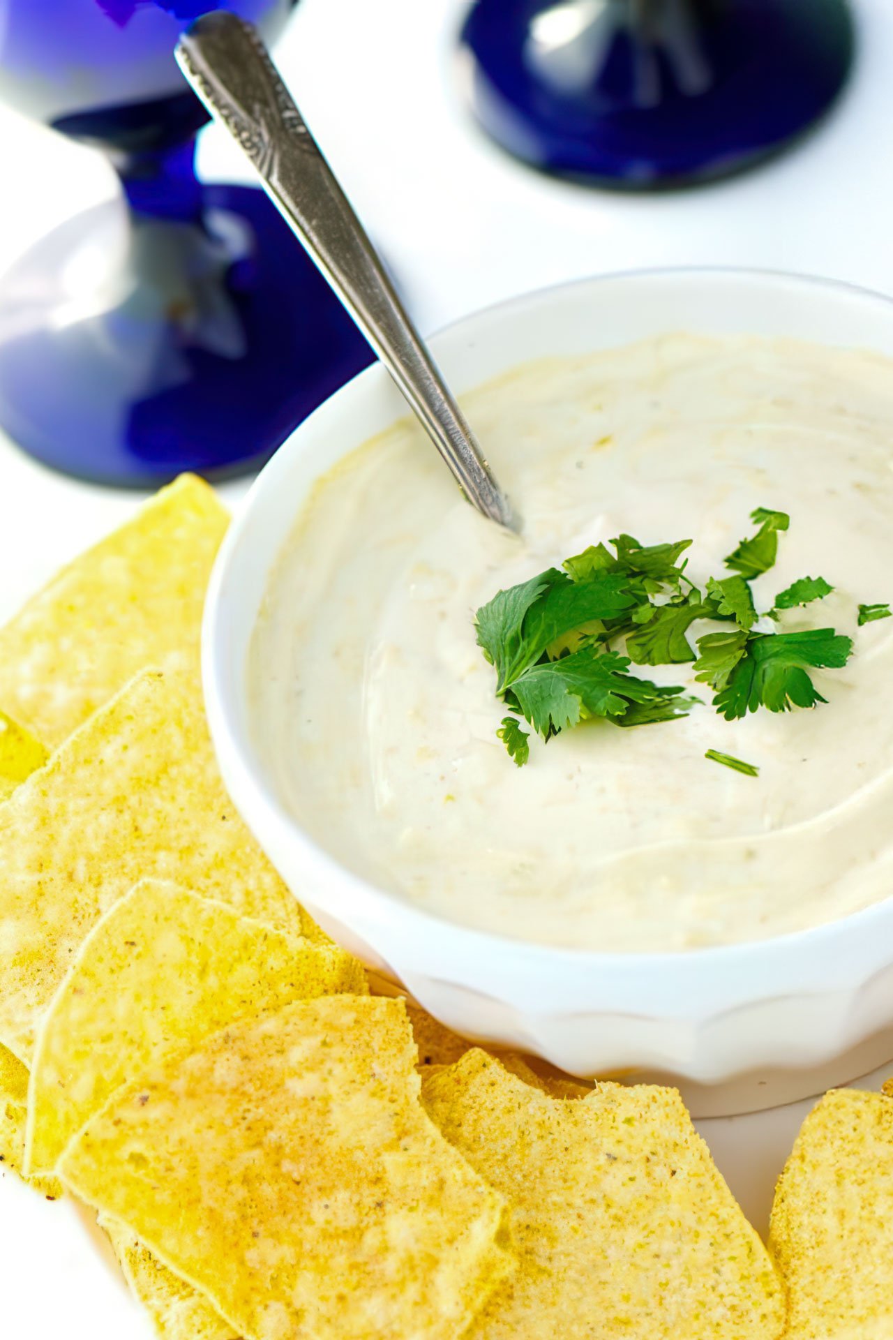 A bowl holds vegan queso dip with chopped cilantro. It's sitting next to tortilla chips and a bright blue stemmed glass is behind it.