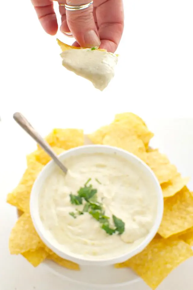 A hand holds a tortilla chip full of vegan queso dip. Below it is the rest of the bowl of the dip and more tortilla chips.