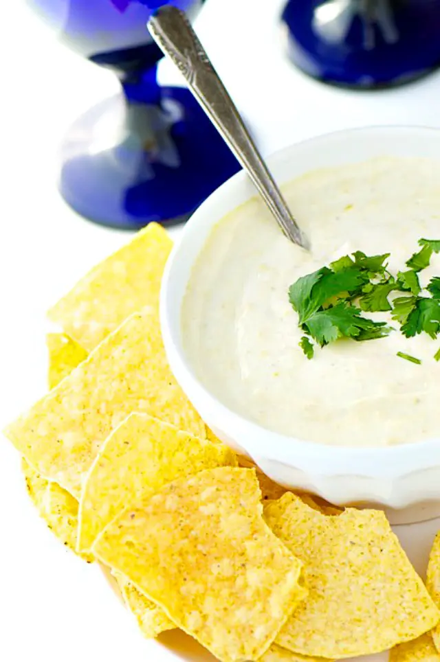 A bowl holds vegan queso dip with chopped cilantro. It's sitting next to tortilla chips and a bright blue stemmed glass is behind it.