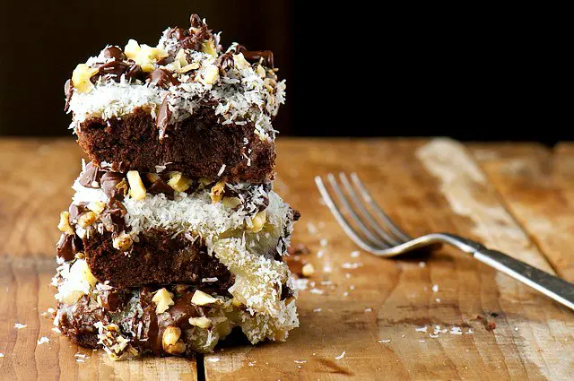 Three magic brownie bars with coconut, chocolate, and a sweet almond milk layer are stacked on a wooden table next to a fork.