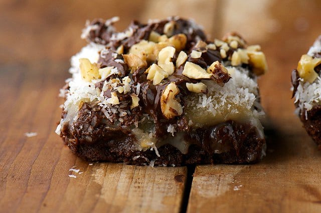 A brownie topped with coconut, chocolate chips and nuts sits on a wooden counter.