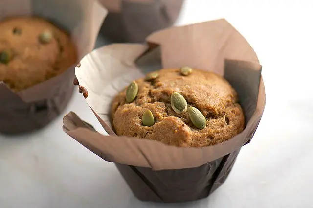 A muffin with pumpkin seeds on top sits in large brown papers on a white counter. There are two other muffins behind it.