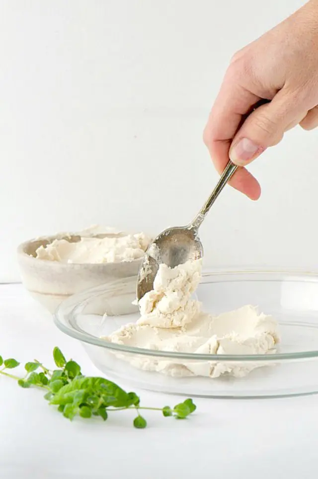 A hand holds a spoon spreading vegan cream cheese into a glass pie pan.