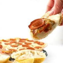 A hand holds crusty bread topped with vegan pizza dip. The container of dip is behind it.