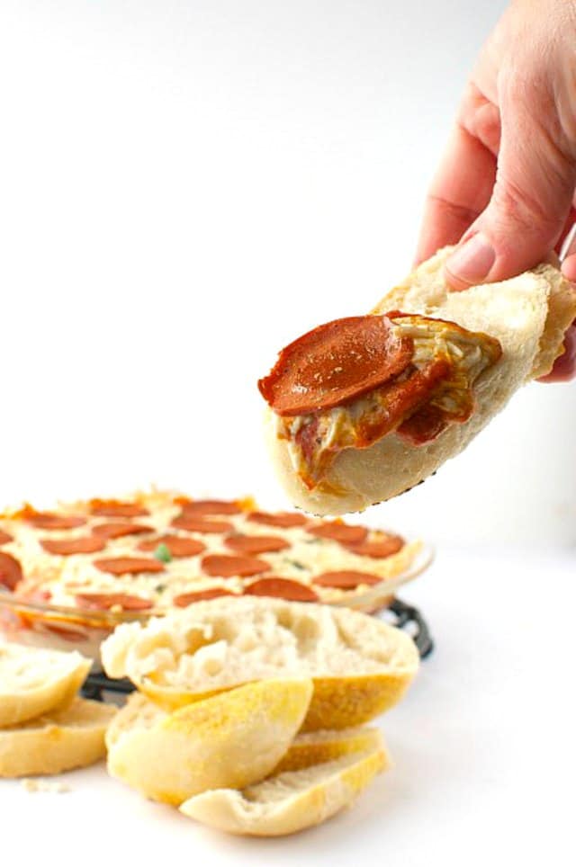A hand holds a piece of bread covered with vegan pepperoni pizza dip. More bread slices and the rest of the dip is behind it.