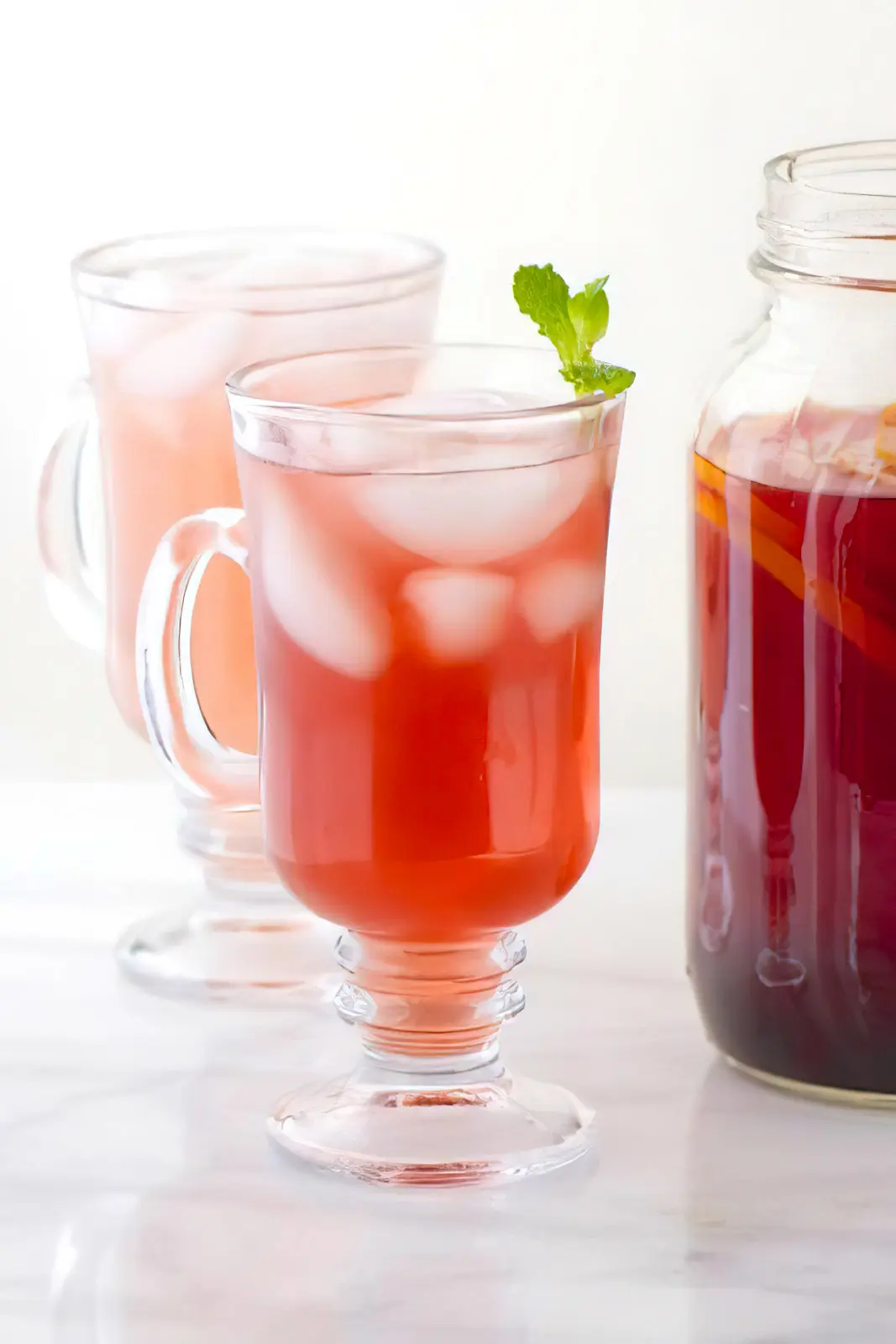 Two tall glasses with ice and punch inside, one with a mint sprig at the top are sitting next to a mason jar full of punch.