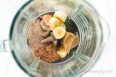 Looking into a blender jar there are ingredients like banana slices, chocolate protein powder, peanut butter, and more.