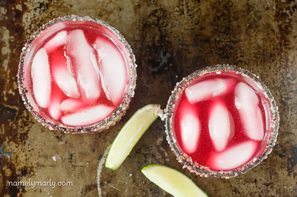 Looking down on two glasses of Hibiscus Margaritas with ice cubes and lime wedges.