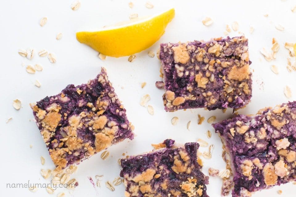 Several slices of blueberry pie bars sit on a white counter next to a lemon slice.