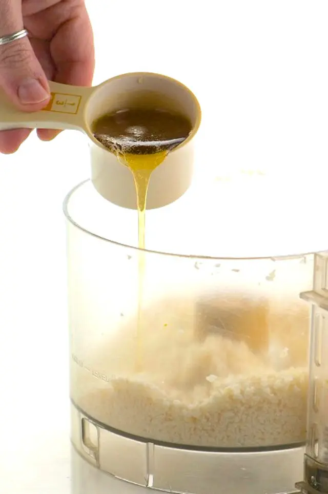 A hand pours syrup into a food processor bowl with coconut shreds.