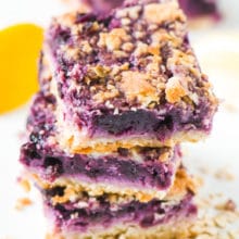 A stack of three vegan blueberry lemon pie bars are surrounded by oatmeal, lemons, and more slices in the background.