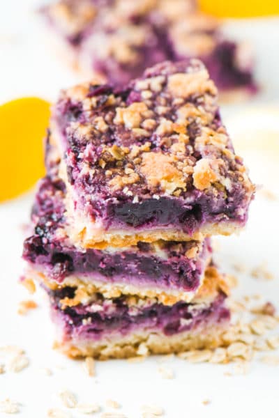 A stack of three vegan blueberry lemon pie bars are surrounded by oatmeal, lemons, and more slices in the background.