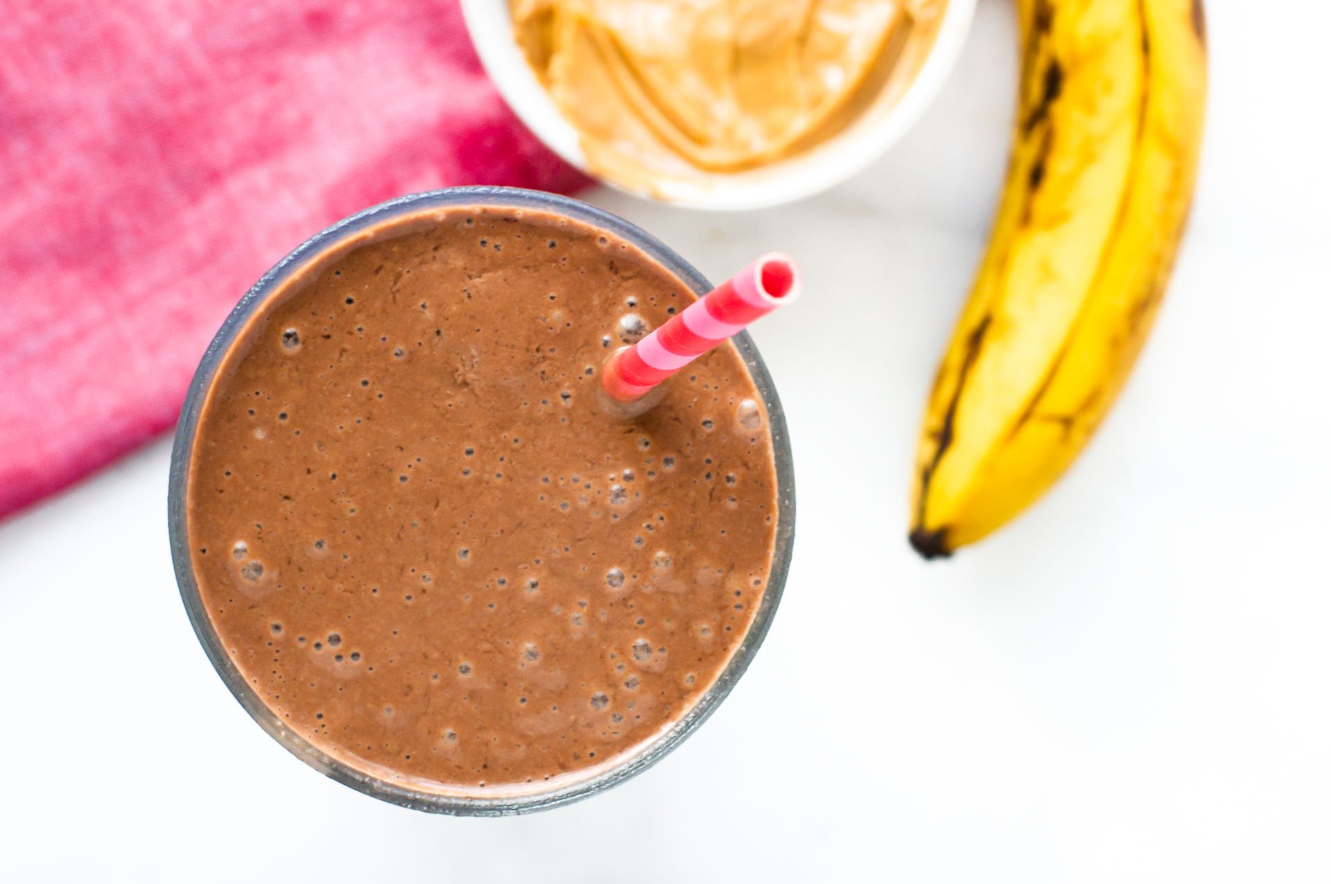 A top-down shot looking at chocolate peanut butter protein in a glass. There's a pink straw in the smoothie. There's a bowl of peanut butter and a ripe banana sitting next to it.