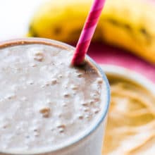 A closeup of a Chocolate Peanut Butter Protein Smoothie in a tall glass with a pink straw. A banana and bowl of peanut butter is blurred in the back.