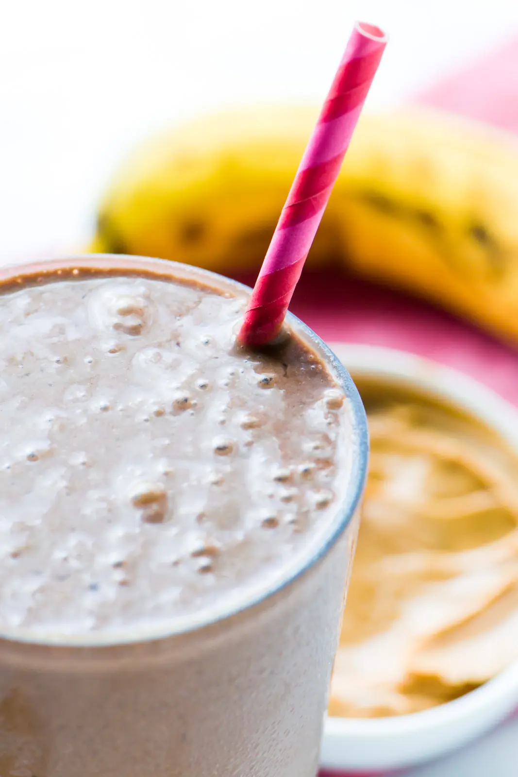A closeup of a chocolate peanut butter smoothie in a glass with a pink striped paper straw. There is peanut butter and a banana behind it.