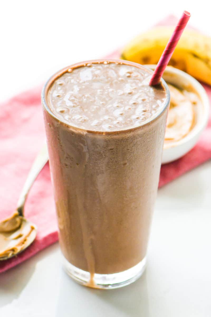 a Chocolate Peanut Butter Protein Smoothie is in a tall glass with a pink straw. A spoon with peanut butter, along with another bowl of peanut butter and a ripe banana sits behind it.
