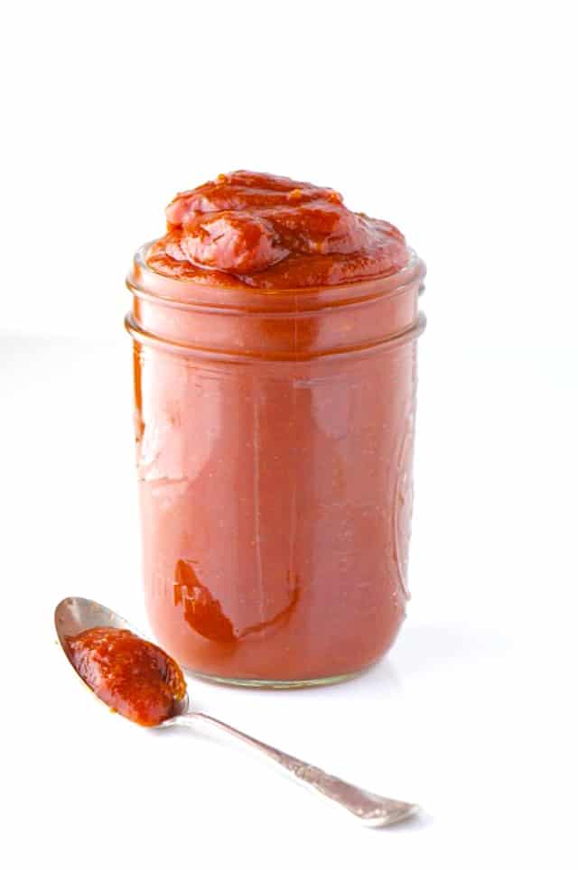 A small mason jar holds date sweetened BBQ sauce. A spoon with more of the sauce is beside it.