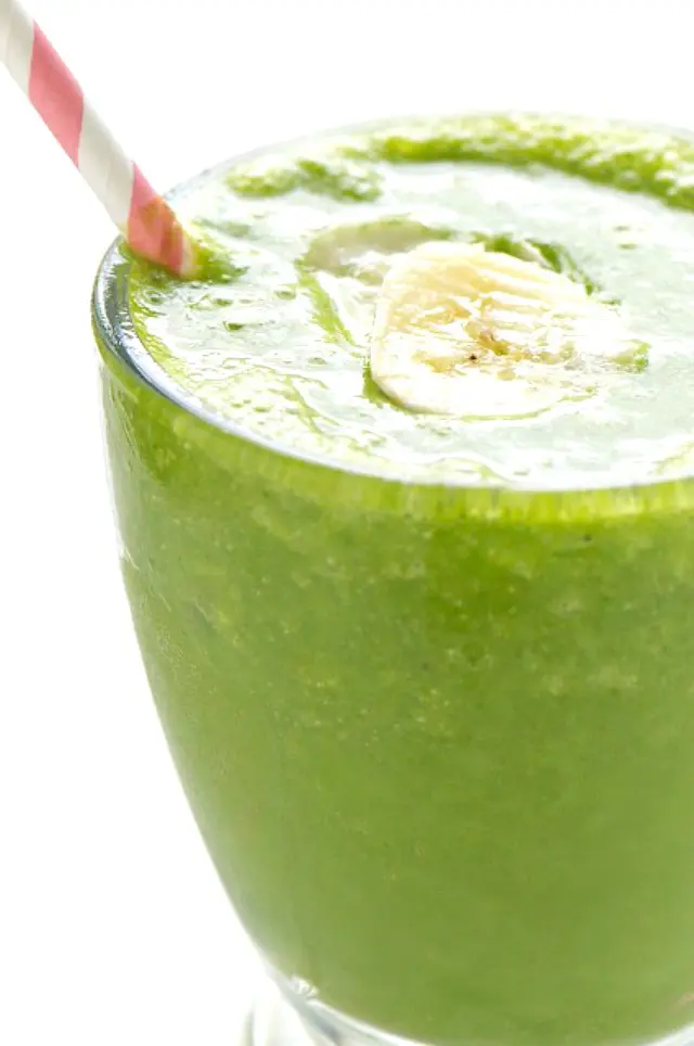 A a clear glass is full to the rim with green banana smoothie. A pink and white paper straw is on the side, and a slice of banana is in the middle of the smoothie.