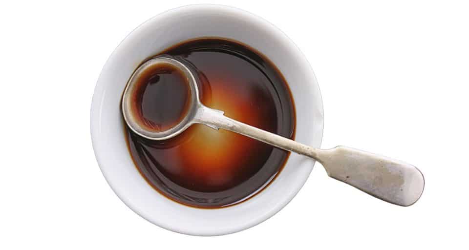 Looking down on a white bowl on a white counter. There is a dark sauce in the bowl and a measuring spoon holds some of the dark sauce.