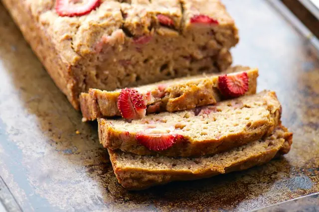 A loaf of quick bread with strawberries on top on a baking sheet with several slices cut off the end.