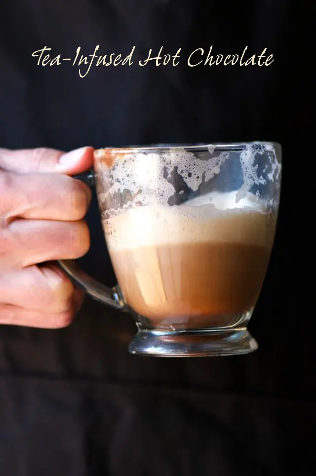 A hand holds a mug of half-full cocoa with frothy milk on the top.