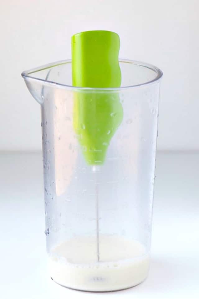 A green frothing device is in a tall container. There is plant-based milk at the bottom.