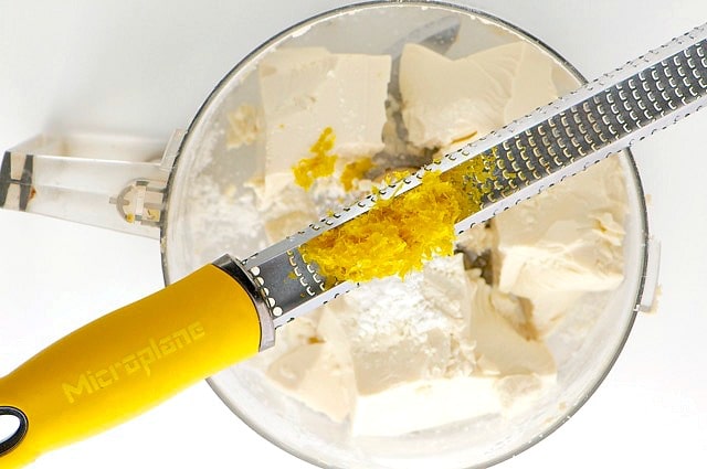 A zester with lemon zest sits on top of a food processor bowl with silken tofu in it.