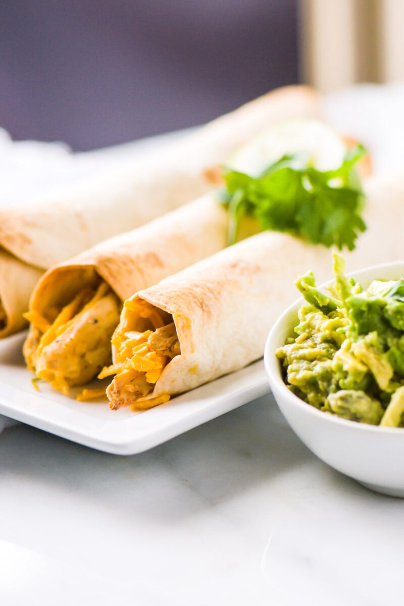 Vegan chicken taquitos are on a plate next to guacamole.