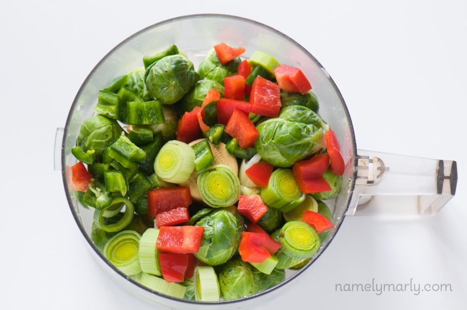 Looking down on a food processor full of raw Brussels sprouts, red bell peppers, and sliced jalape&ntilde;os. 