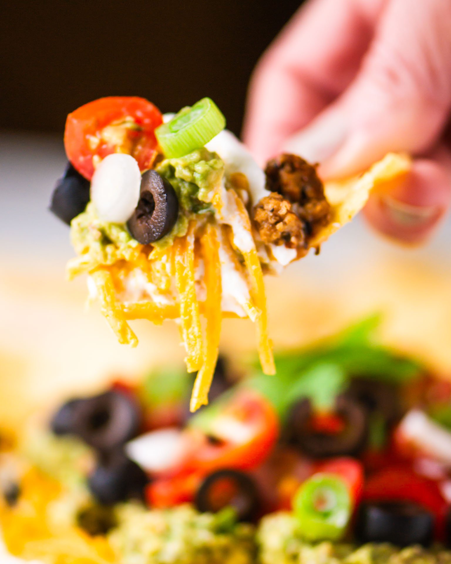 A hand holds a tortilla chip loaded with vegan seven layer dip. The plate of dip is sitting in the background.
