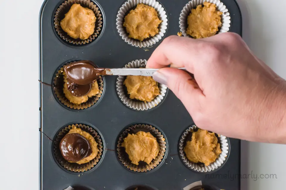 A hand holds a spoon and is dropping melted chocolate over peanut butter cups in a mini muffin pan.
