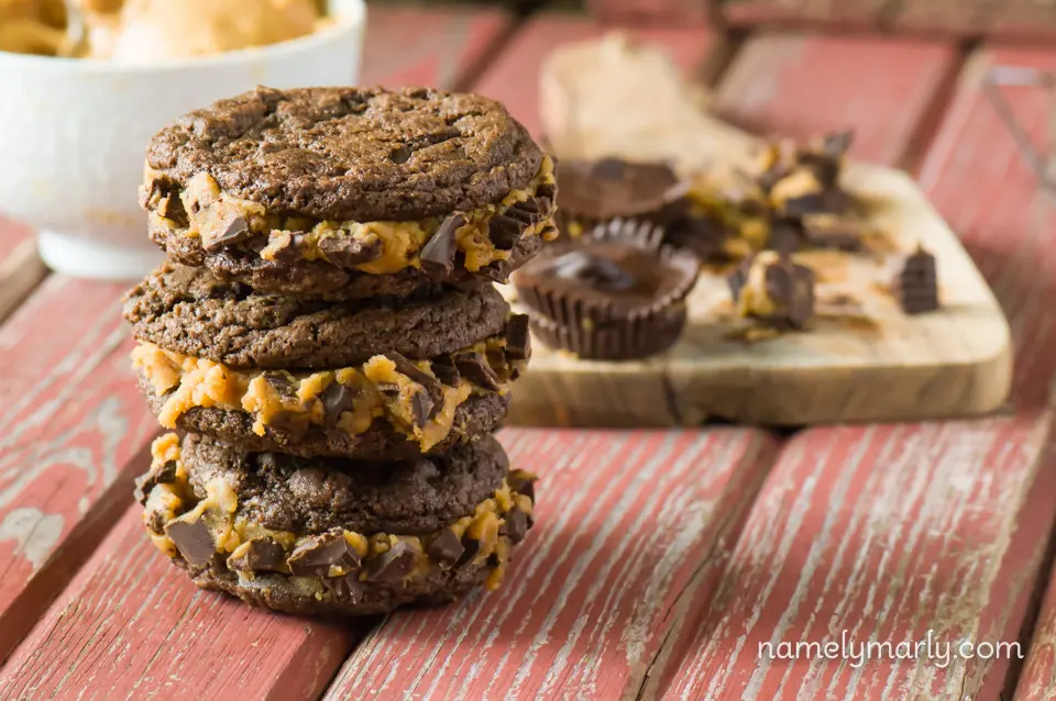 A stack of Chocolate Peanut Butter Cup Sandwich Cookies sits in front of vegan peanut butter cups and a bowl of peanut butter.