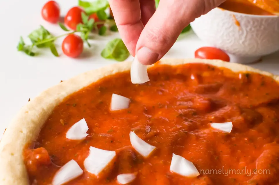 A hand is placing chopped onion on a pizza crust topped with red sauce. There are basil leaves and cherry tomatoes in the background.