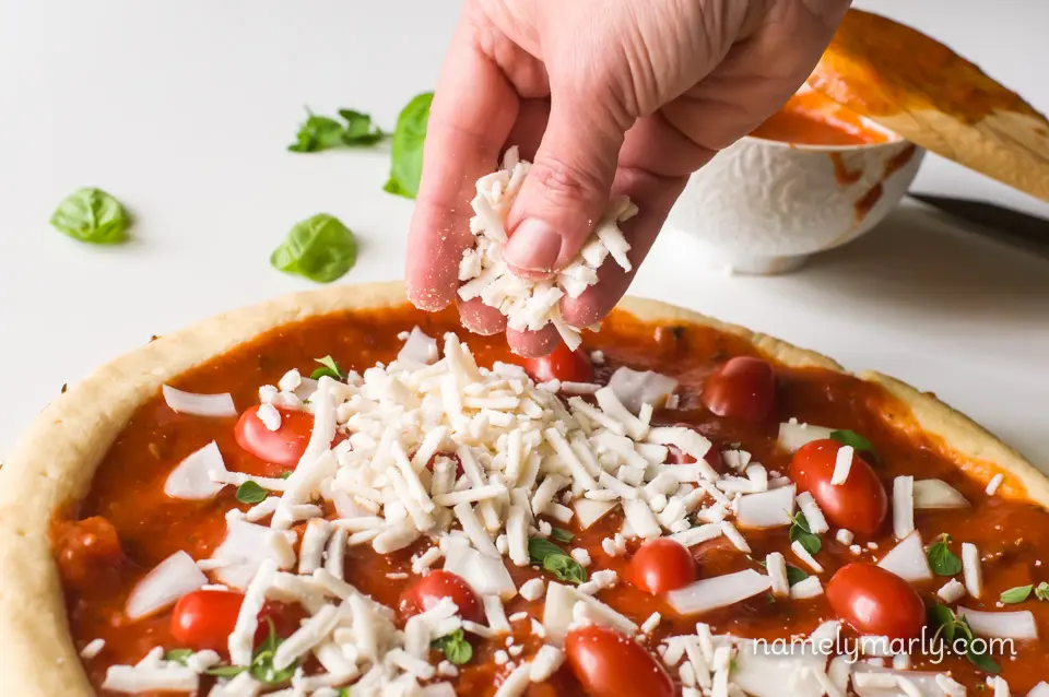 A hand is spreading mozzarella shreds over a pizza. There are basil leaves in the background.